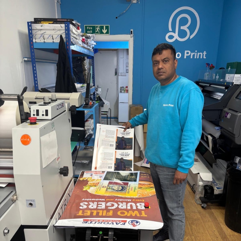 A print shop owner in Forest Gate, East London, holds a print sheet by a laminating machine over a burger menu print